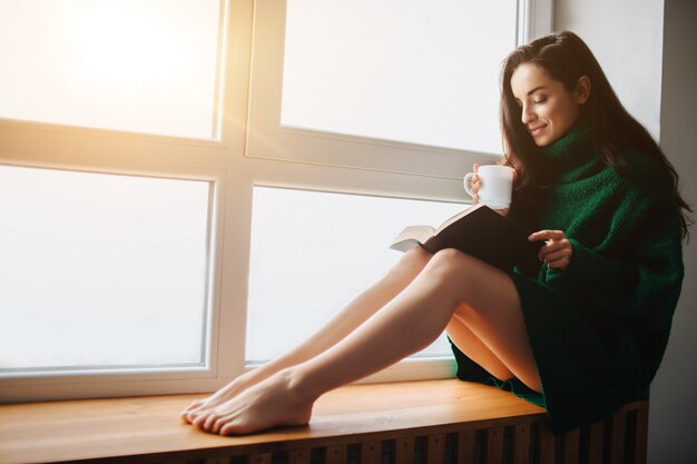 Une jeune femme brune est assise sur un rebord de fenêtre et tenant un livre et une tasse de thé à la main