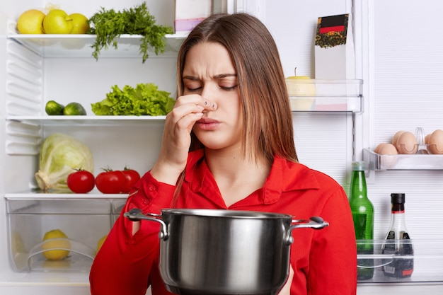 Jeune femme brune avec une expression mécontente sent la soupe gâtée dans un ragoût, sent l'odeur de moisi dans la cuisine à domicile, se dresse contre le réfrigérateur.