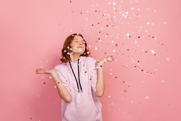 Jeune femme brune extrêmement heureuse appréciant la chute de confettis en forme de coeur et souriant les yeux fermés, célébrant la Saint-Valentin, l'ambiance des vacances. Tourné en studio intérieur, isolé sur fond rose