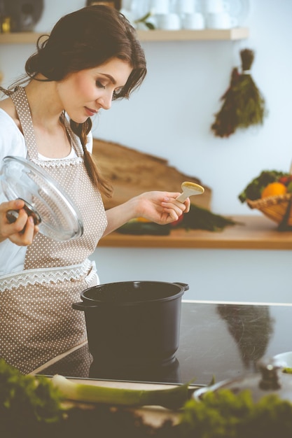 Jeune femme brune faisant cuire la soupe dans la cuisine. Femme au foyer tenant une cuillère en bois dans sa main. Concept de nourriture et de santé.