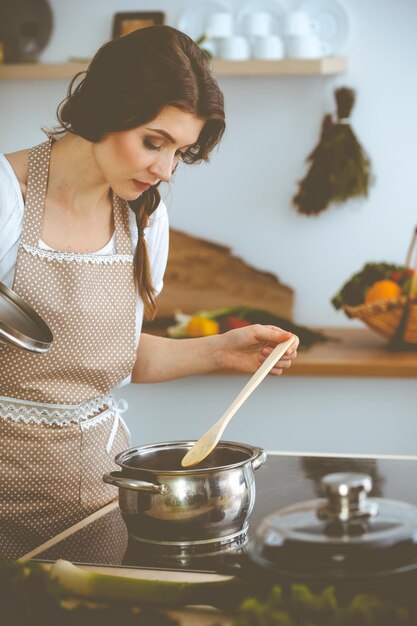 Jeune femme brune faisant cuire la soupe dans la cuisine. Femme au foyer tenant une cuillère en bois dans sa main. Concept de nourriture et de santé.