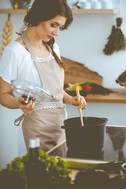 Jeune femme brune faisant cuire la soupe dans la cuisine. Femme au foyer tenant une cuillère en bois dans sa main. Concept de nourriture et de santé.