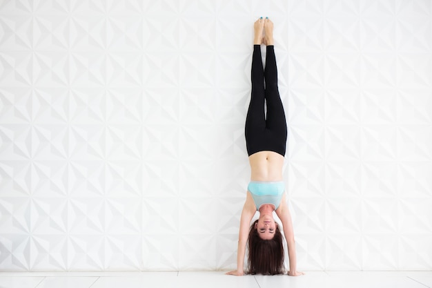 Jeune femme brune faisant le poirier sur un mur blanc