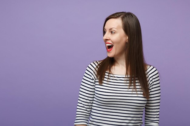 Jeune femme brune fille dans des vêtements rayés décontractés posant isolé sur fond de mur violet violet portrait en studio. Concept de mode de vie des émotions sincères des gens. Maquette de l'espace de copie En regardant de côté le sourire.