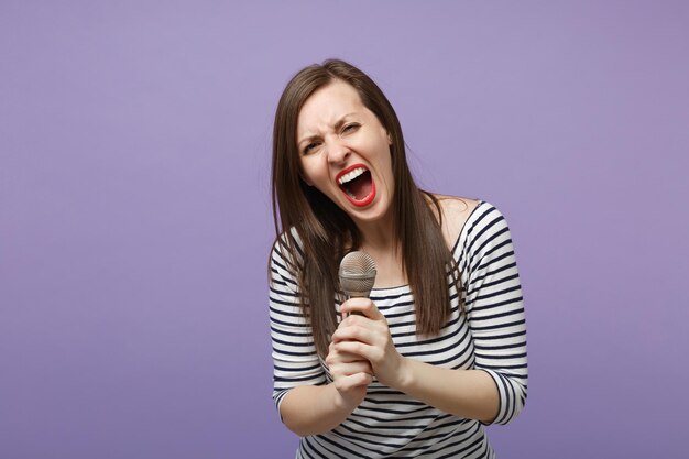 Jeune femme brune fille dans des vêtements rayés décontractés posant isolé sur fond violet violet portrait en studio. Concept de mode de vie des émotions sincères des gens. Maquette de l'espace de copie. Chantez la chanson dans le microphone.