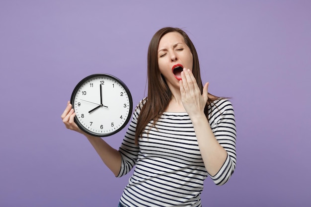 Jeune femme brune fille dans des vêtements rayés décontractés posant isolé sur fond violet violet portrait en studio. Concept de mode de vie des émotions sincères des gens. Maquette de l'espace de copie. Horloge Ronde Tenir dans la main