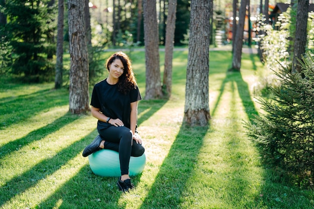 Une jeune femme brune en forme se repose après avoir fait des exercices au ballon de fitness mène un style de vie sportif actif pose en milieu urbain sur l'herbe verte contre des arbres avec du soleil fait de la gymnastique