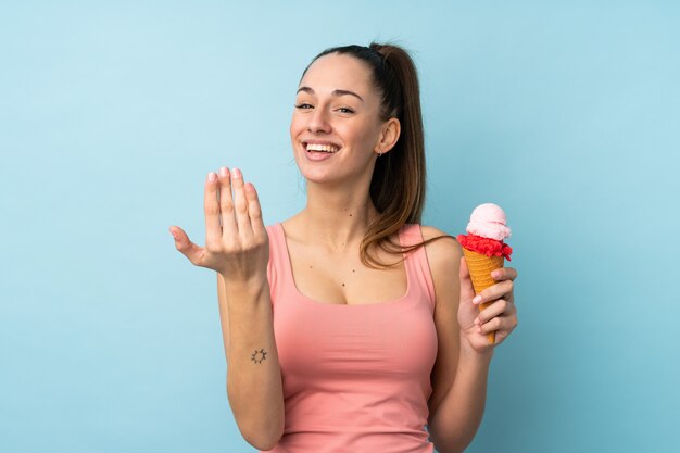 Jeune femme brune avec une glace au cornet sur un mur bleu isolé invitant à venir avec la main.