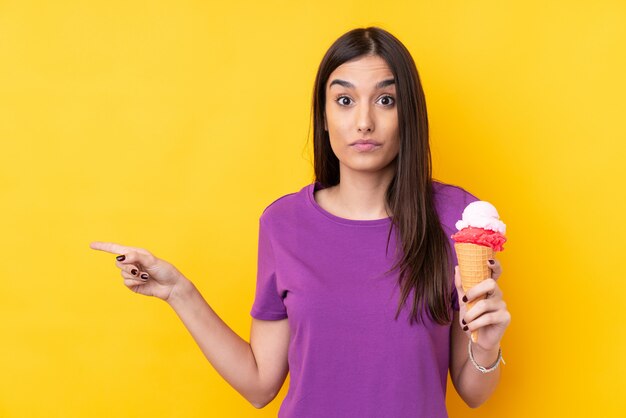 Jeune femme brune avec une glace au cornet sur un mur jaune isolé surpris et pointant le côté