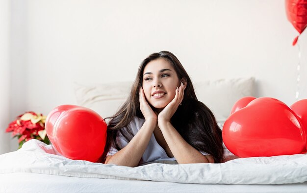 Jeune femme brune heureuse pose dans le lit avec des ballons en forme de coeur rouge