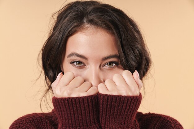 Jeune femme brune heureuse et positive posant isolée sur un mur beige couvrant le visage avec les mains.