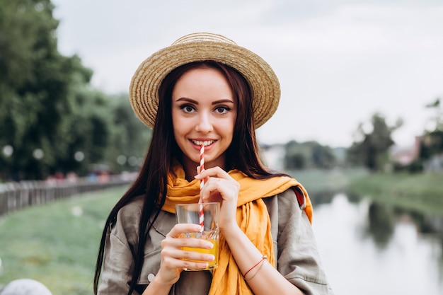 Jeune femme brune hipster, boire du jus d'orange à l'extérieur, souriant, profitant de ses vacances. Vacances d'été dans la ville. Closeup portrait d'une jeune fille dans un chapeau élégant dans le parc.