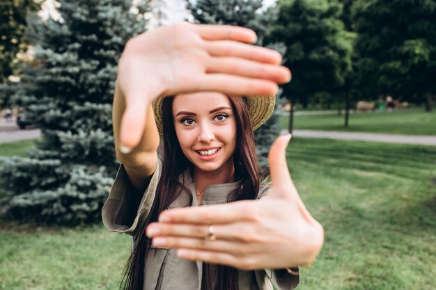 Jeune femme brune hipster prenant un selfie, montrant la langue, la grimace posant à l'extérieur. Closeup portrait d'une jeune fille dans un chapeau d'été élégant dans un parc.