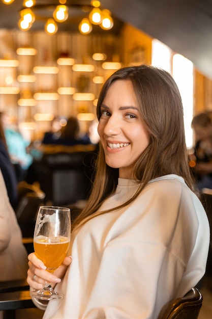 Une jeune femme brune joyeuse qui te regarde en faisant un toast avec un verre de bière.