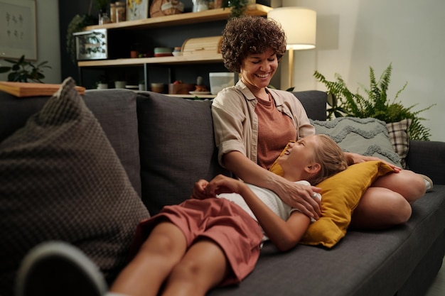 Jeune femme brune joyeuse en tenue décontractée assise sur un canapé avec sa fille