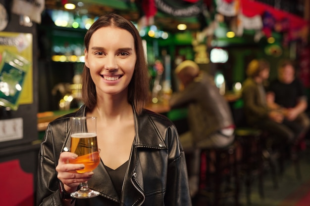 Jeune Femme Brune Joyeuse Vous Regarde Tout En Grillant Avec Un Verre De Bière Devant La Caméra Contre Des Gars Assis Au Comptoir Du Bar Dans Un Pub