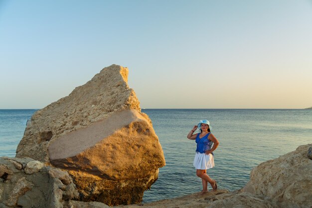 Une jeune femme brune en jupe blanche et un chapeau de soleil au bord de la mer sur un rocher admire la nature.
