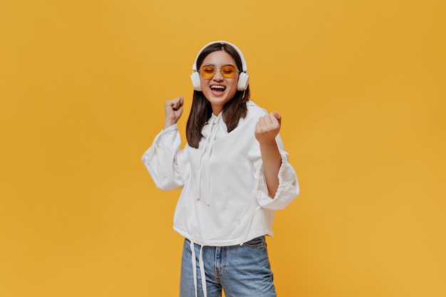 Jeune femme brune à lunettes de soleil orange, sweat à capuche blanc et jeans chante et écoute de la musique dans les écouteurs sur le mur orange