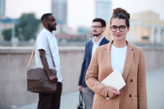 Jeune femme brune manager avec tablette numérique debout devant la caméra en milieu urbain contre deux hommes d'affaires parlant