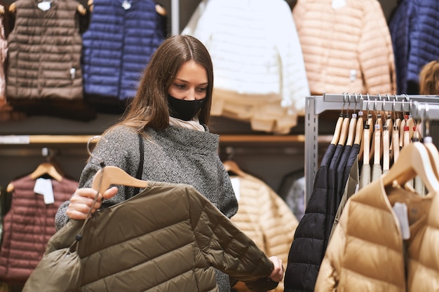 Jeune femme brune en masque médical choisit des vêtements dans le magasin et fait des achats