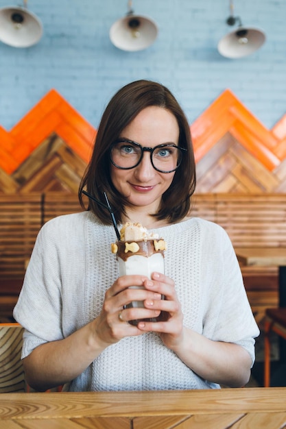 Jeune femme brune mignonne boit un milk-shake joliment décoré avec de la crème et des biscuits dans le café. Tonifiant.