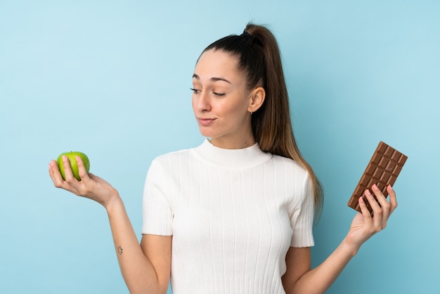 Jeune femme brune sur mur bleu isolé ayant des doutes tout en prenant une tablette de chocolat dans une main et une pomme dans l'autre