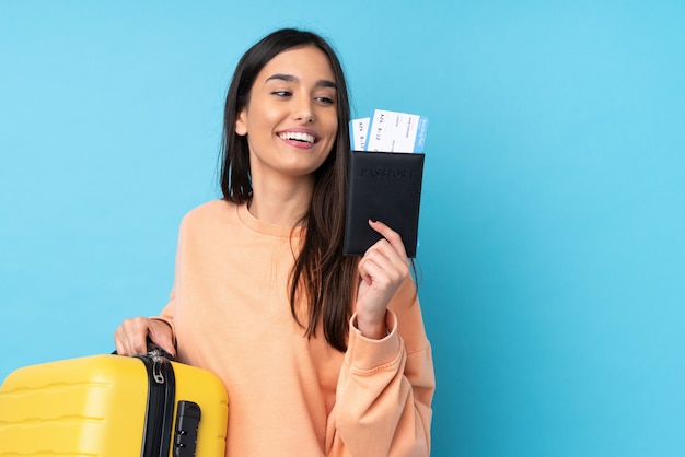 Jeune femme brune sur mur bleu isolé en vacances avec valise et passeport