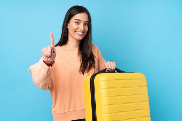 Jeune femme brune sur mur bleu isolé en vacances avec valise de voyage et en compter un