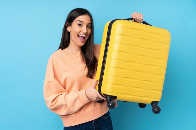 Jeune femme brune sur mur bleu isolé en vacances avec valise de voyage