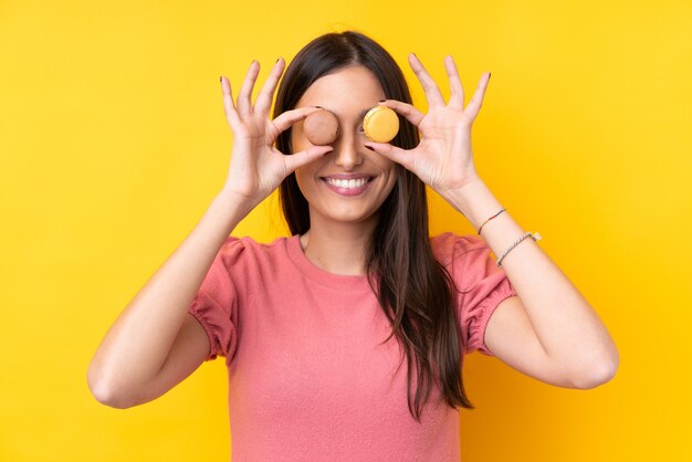 Jeune femme brune sur un mur jaune isolé portant des macarons français colorés comme lunettes
