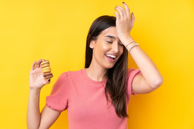 Jeune femme brune sur un mur jaune isolé tenant des macarons français colorés et envisageant la solution