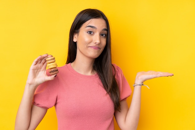 Jeune femme brune sur un mur jaune isolé tenant des macarons français colorés et faisant des doutes geste