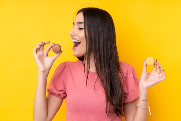 Jeune femme brune sur un mur jaune isolé tenant des macarons français colorés et le manger