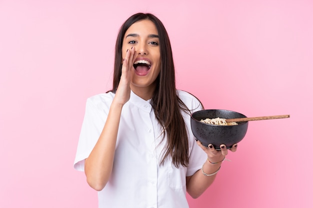 Jeune femme brune sur mur rose isolé criant avec la bouche grande ouverte tout en tenant un bol de nouilles avec des baguettes