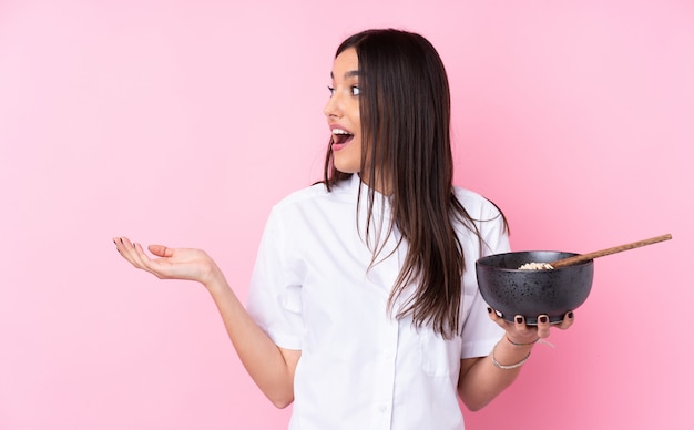 Jeune femme brune sur un mur rose isolé avec une expression faciale surprise tout en tenant un bol de nouilles avec des baguettes