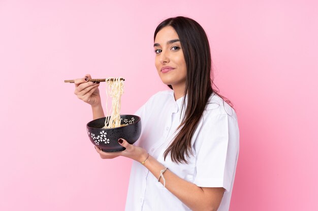 Jeune femme brune sur un mur rose isolé tenant un bol de nouilles avec des baguettes