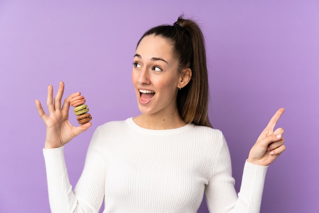 Jeune femme brune sur un mur violet isolé tenant des macarons français colorés et ayant l'intention de réaliser la solution