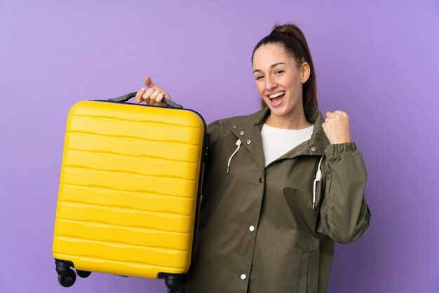 Jeune femme brune sur mur violet isolé en vacances avec valise de voyage