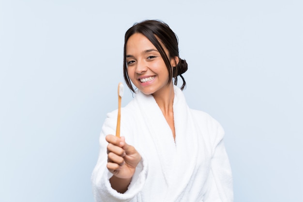 Jeune femme brune en peignoir se brosser les dents sur un mur bleu isolé avec expression heureuse