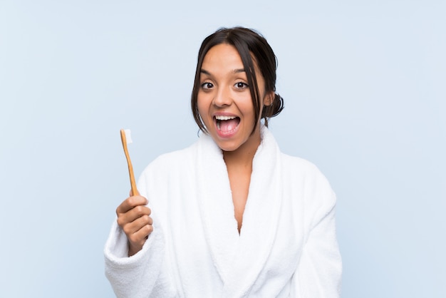 Jeune femme brune en peignoir se brosser les dents sur un mur bleu isolé avec surprise et expression faciale choquée