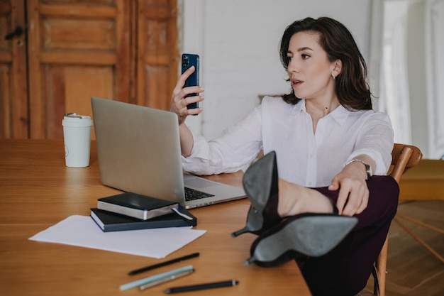 Jeune femme brune perplexe en chemise blanche pantalon sombre tenant le téléphone assis au bureau avec ordinateur portable