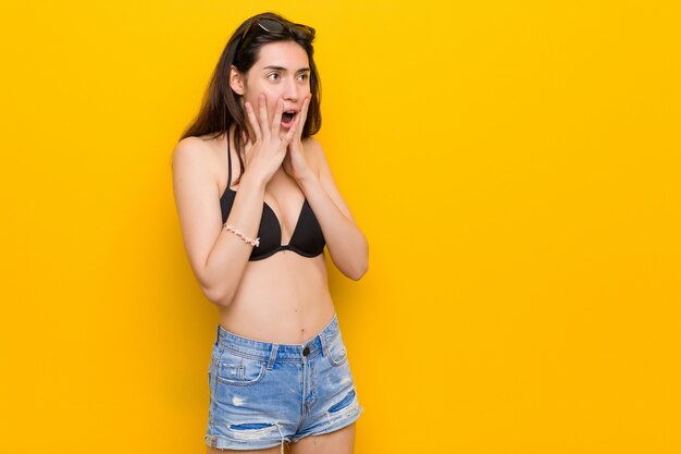 Jeune femme brune portant un bikini contre le mur jaune crie fort, garde les yeux ouverts et les mains tendues.