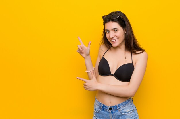 Jeune femme brune portant un bikini contre le mur jaune pointant avec les index vers un espace vide