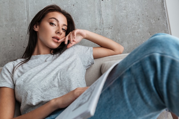 jeune femme brune portant des vêtements décontractés se détendre sur un canapé, tenant un manuel