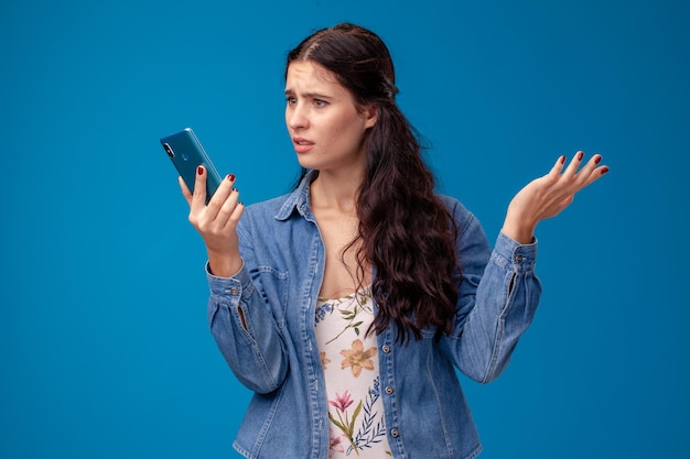 Jeune femme brune pose avec un smartphone debout sur fond bleu.