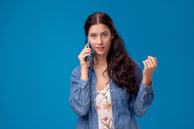 Jeune femme brune pose avec un smartphone debout sur fond bleu.