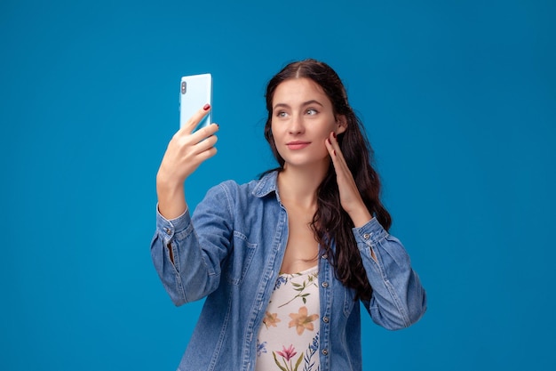 Jeune femme brune pose avec un smartphone debout sur fond bleu.