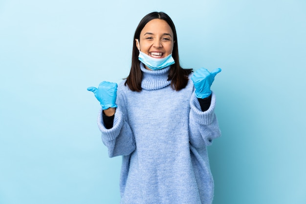 Jeune femme brune de race mixte protégeant contre le coronavirus avec un masque et des gants sur un mur bleu isolé avec les pouces vers le haut de geste et souriant
