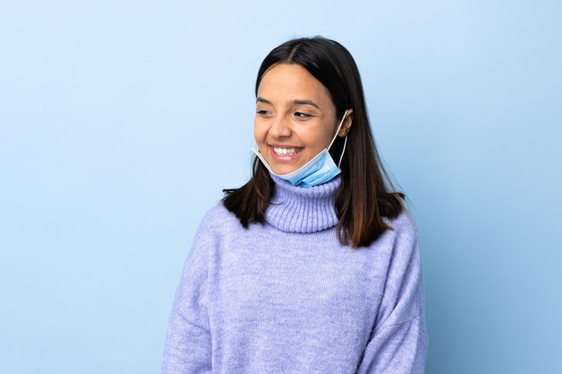 Jeune femme brune de race mixte protégeant contre le coronavirus avec un masque et des gants sur un mur bleu isolé à la recherche sur le côté et souriant