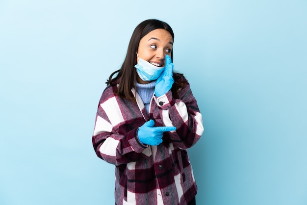 Jeune femme brune de race mixte protégeant du coronavirus avec un masque et des gants sur un mur bleu isolé pointant vers le côté pour présenter un produit et chuchoter quelque chose